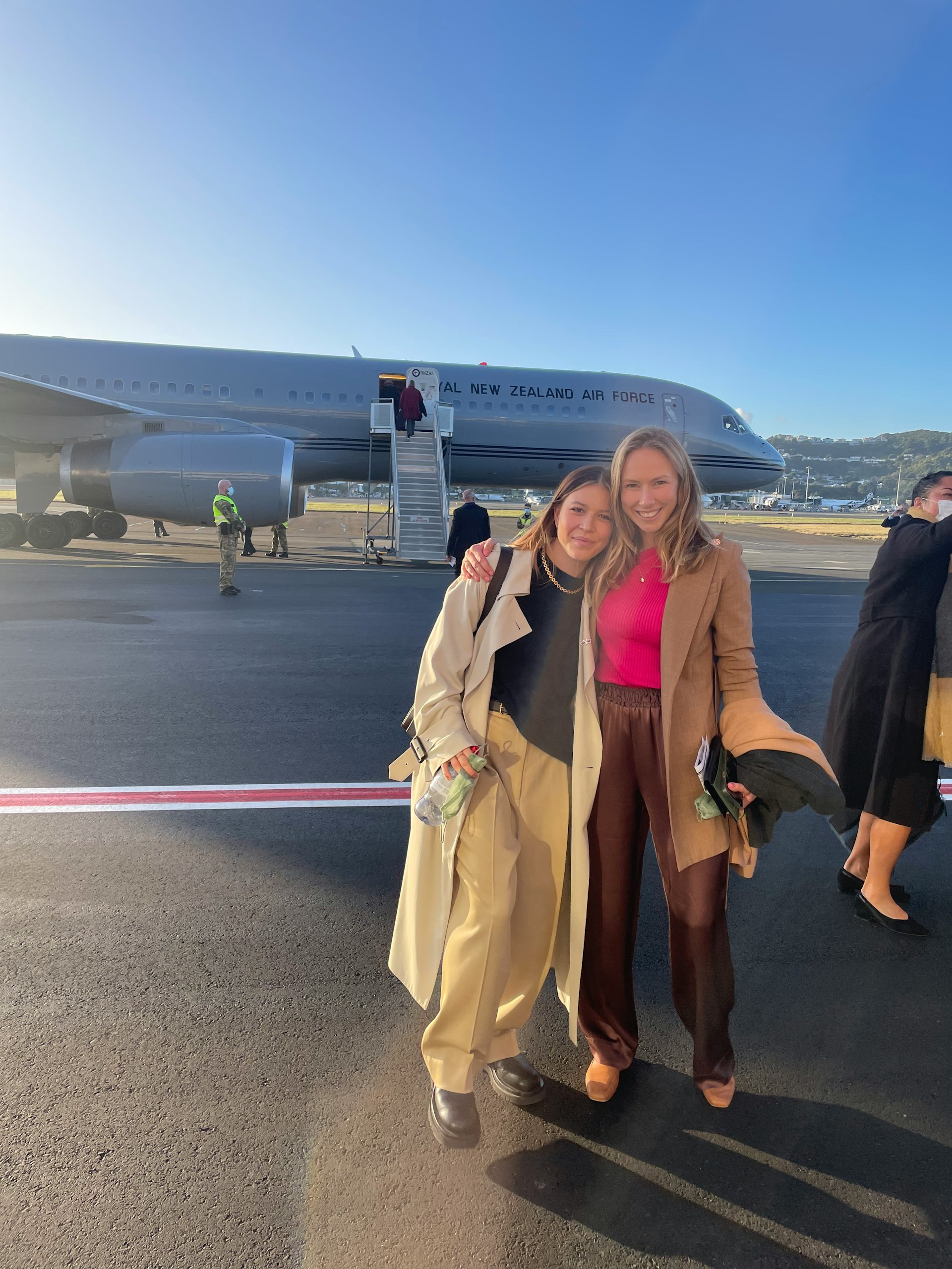 Brooke Roberts and Jessie Wong are smiling into the camera, standing on an airport runway in front of a Royal New Zealand Air Force plane. 