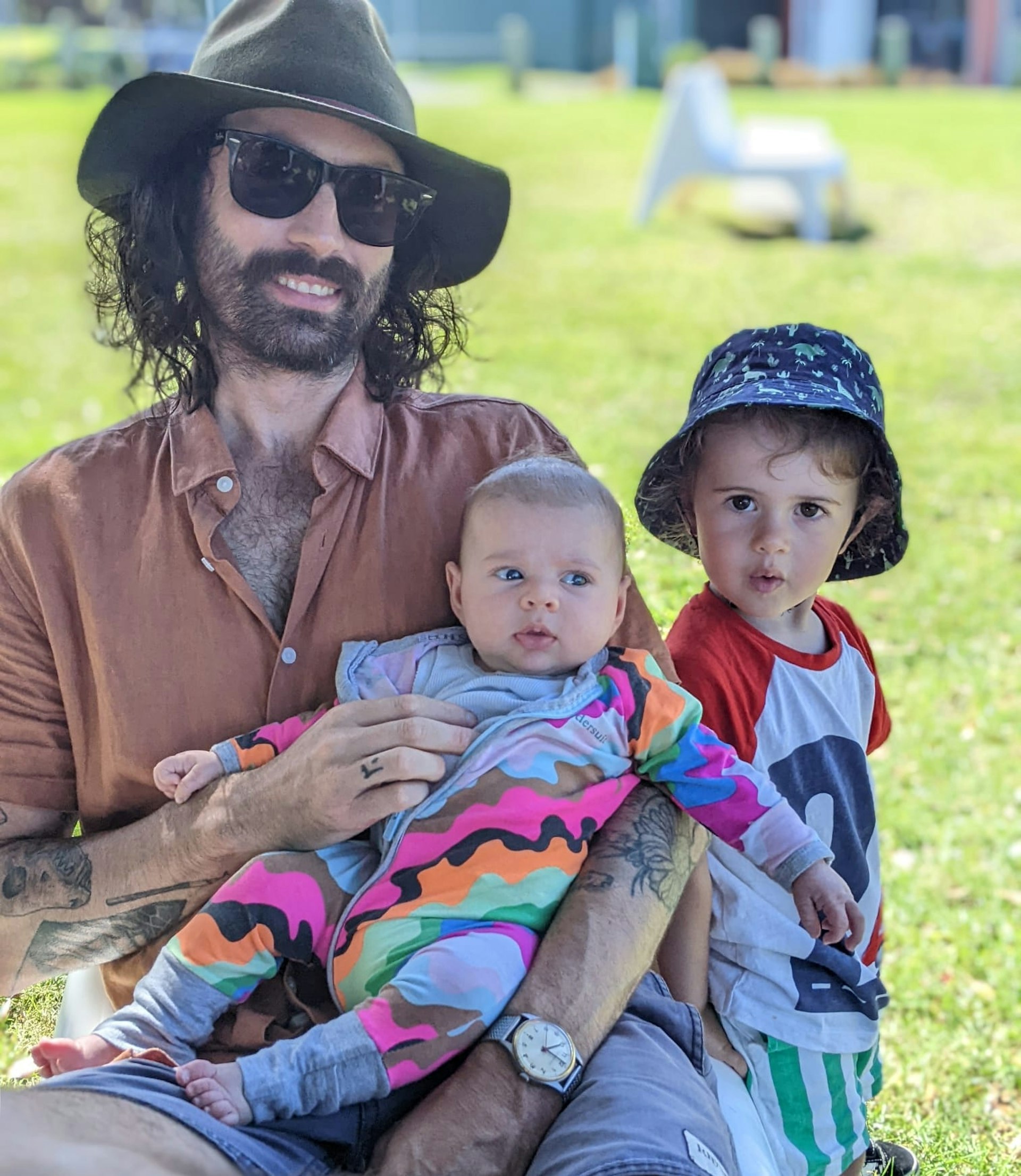 A man wearing a hat and sunglasses sitting down on the grass with his two children