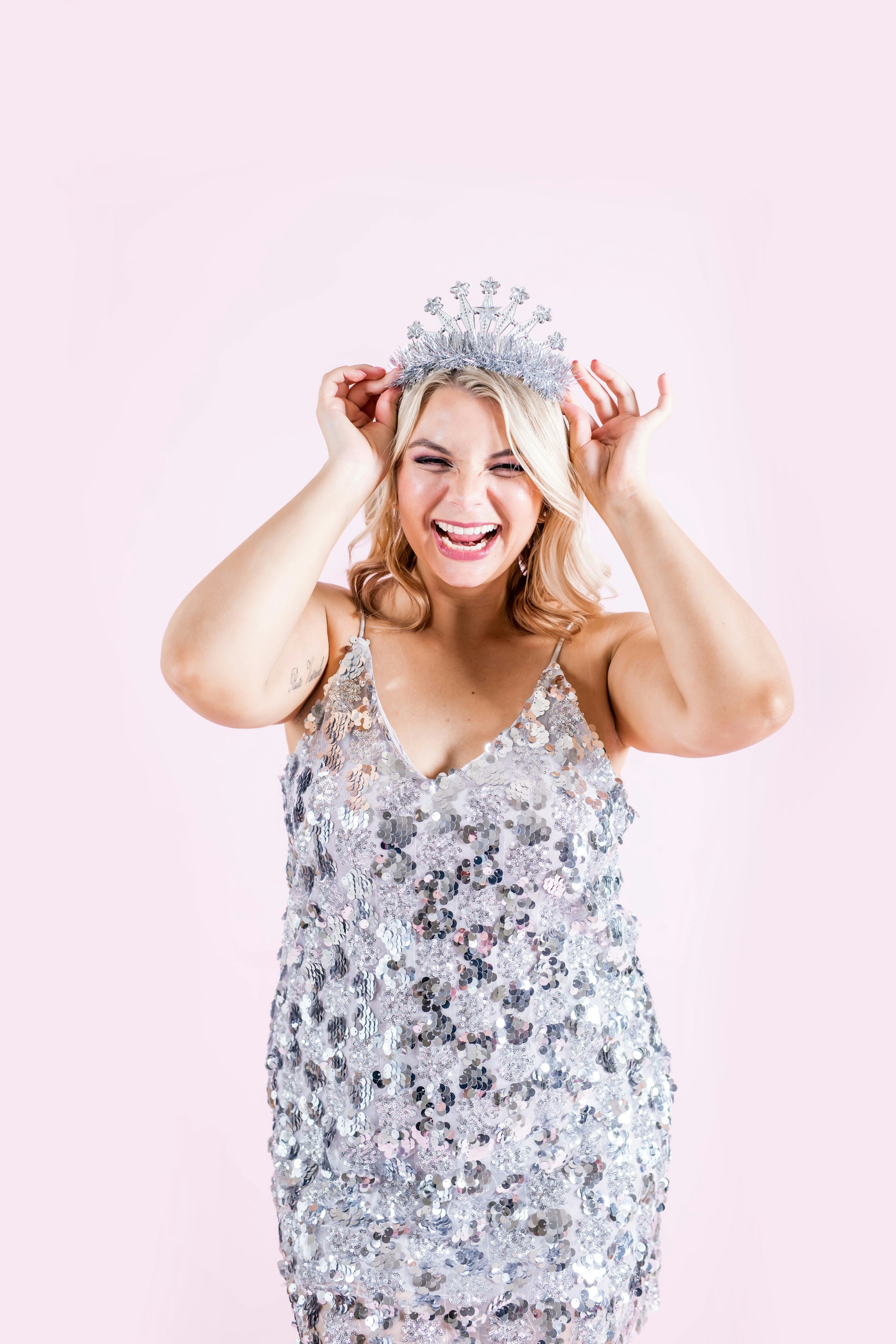 A woman smiling in front of a pink wall wearing a glittery silver dress and holding a crown