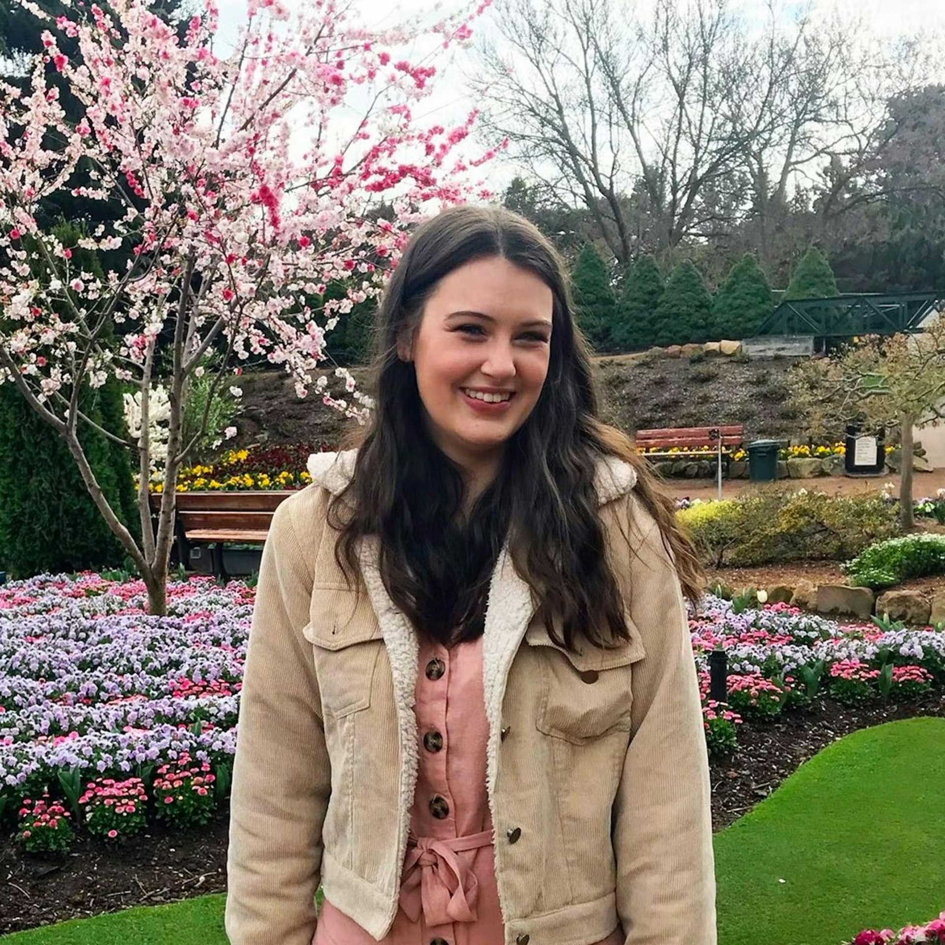 Alyssia is standing in a garden, in front of a Cherry Blossom tree, wearing a pink top and beige jacket and smiling directly into the camera. 