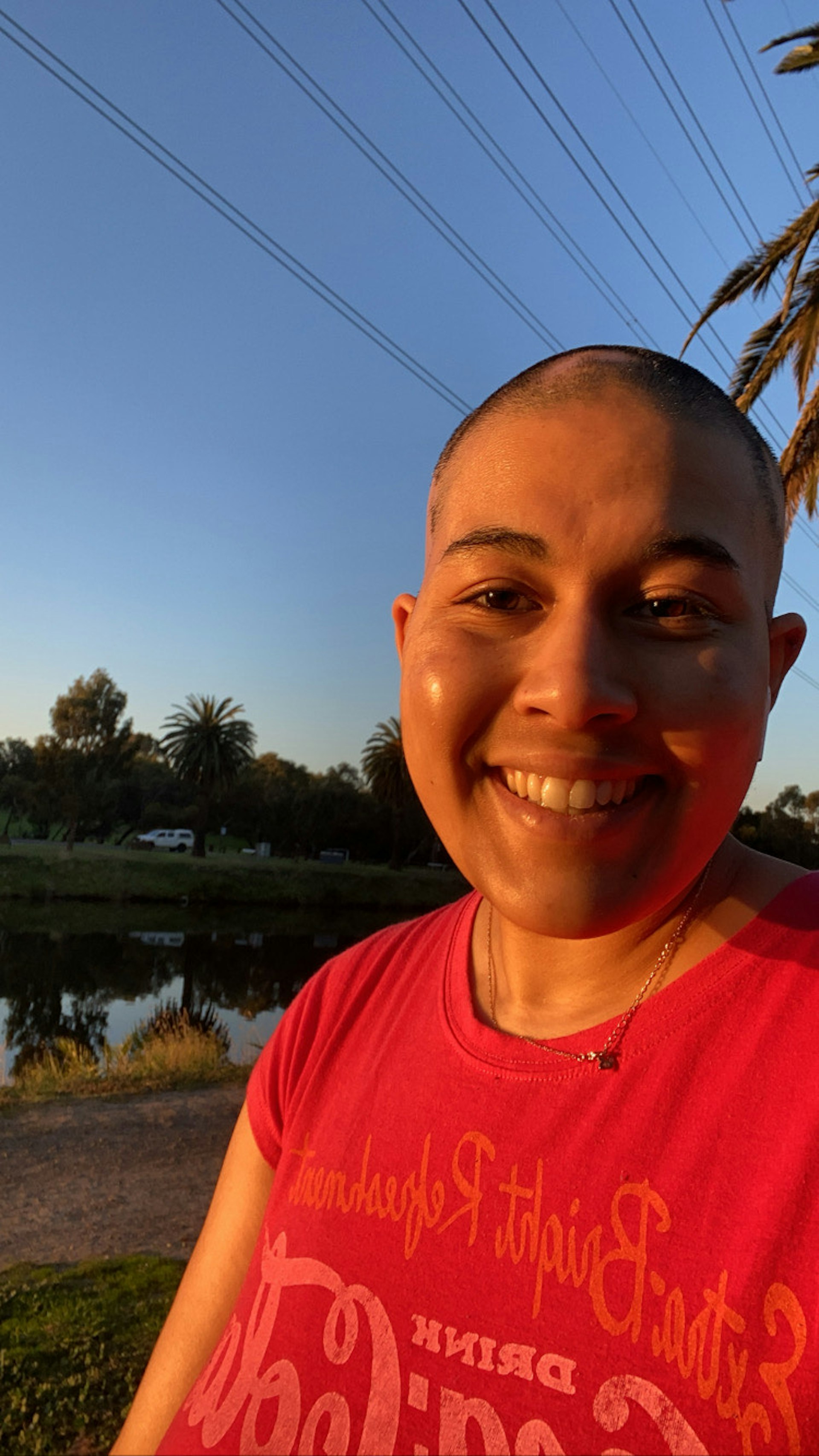Brooke stands outside in front of a clear blue sky, smiling and looking into the camera. She wears a red t-shirt and has short dark hair, and has golden hour sunlight on her face.