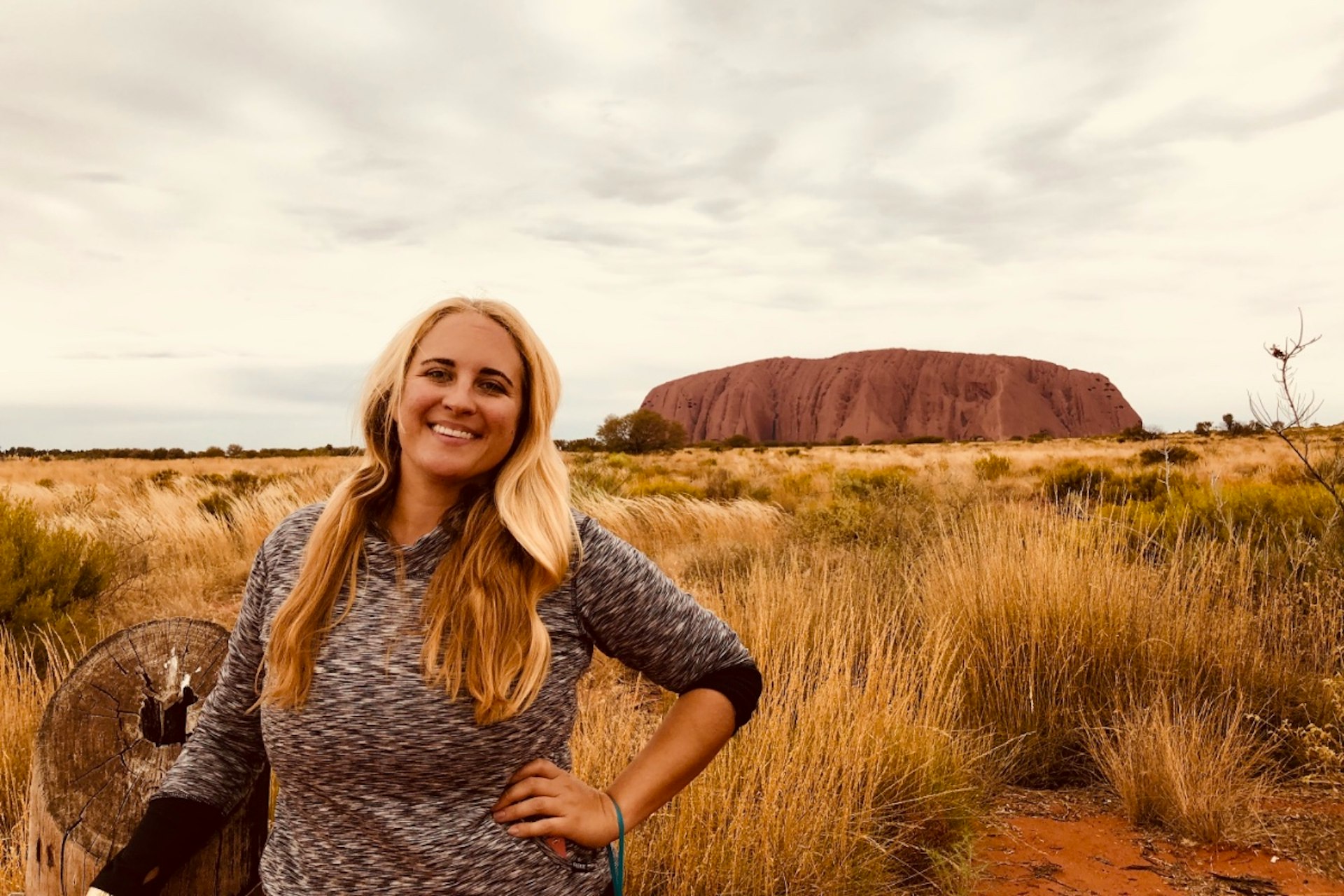 Adrien is standing in front of Uluru in the Australian outback, smiling into the camera with her left hand on her hip. She has long blonde hair, and is wearing a black and white top.