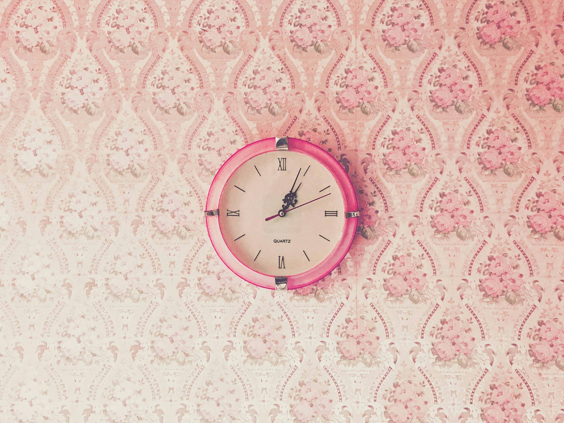 A pink clock with roman numerals hanging on a wall of garish wallpaper.