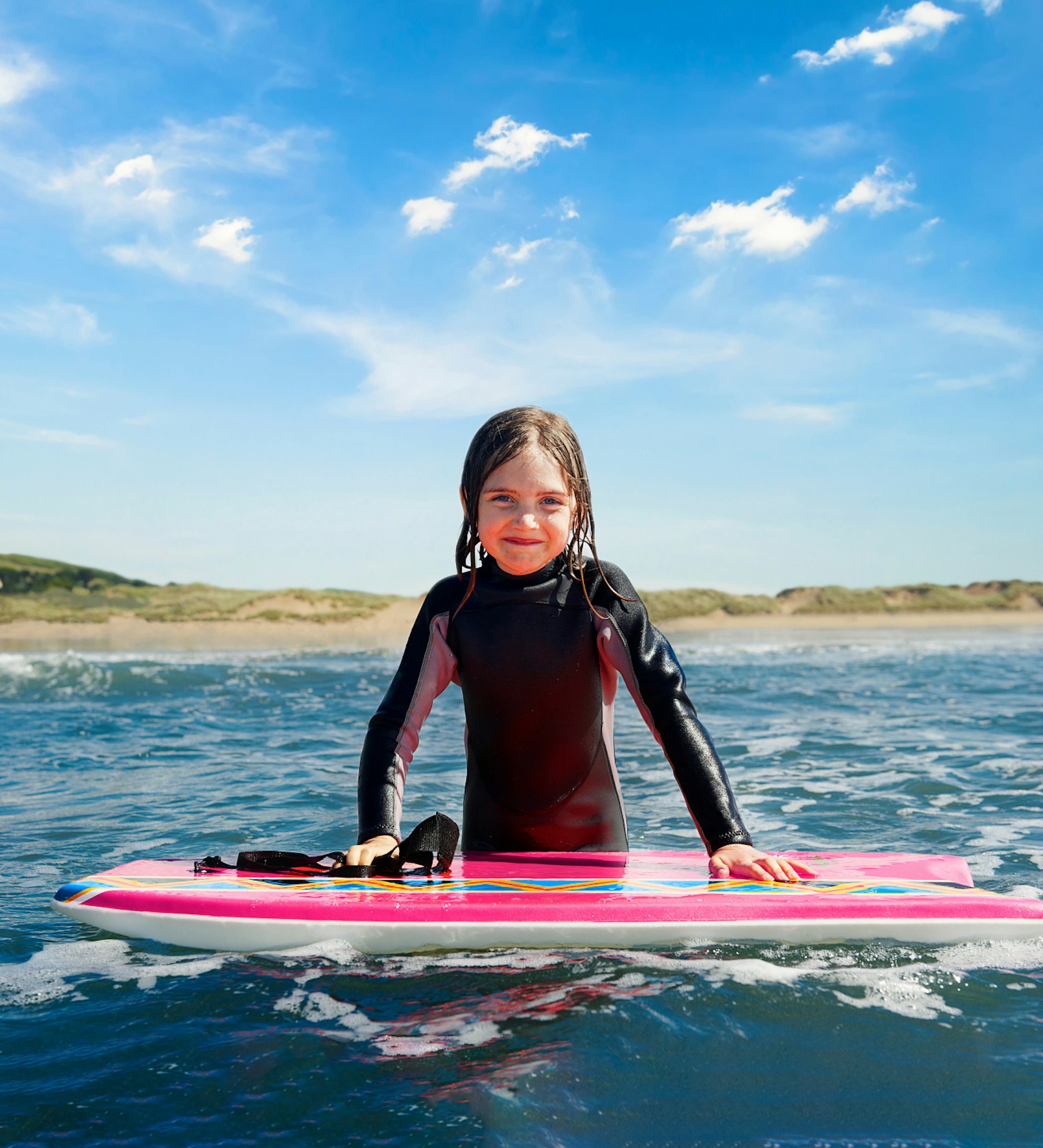 A young girl in a wet suit stands in the sea with her hands on a boogie board.
