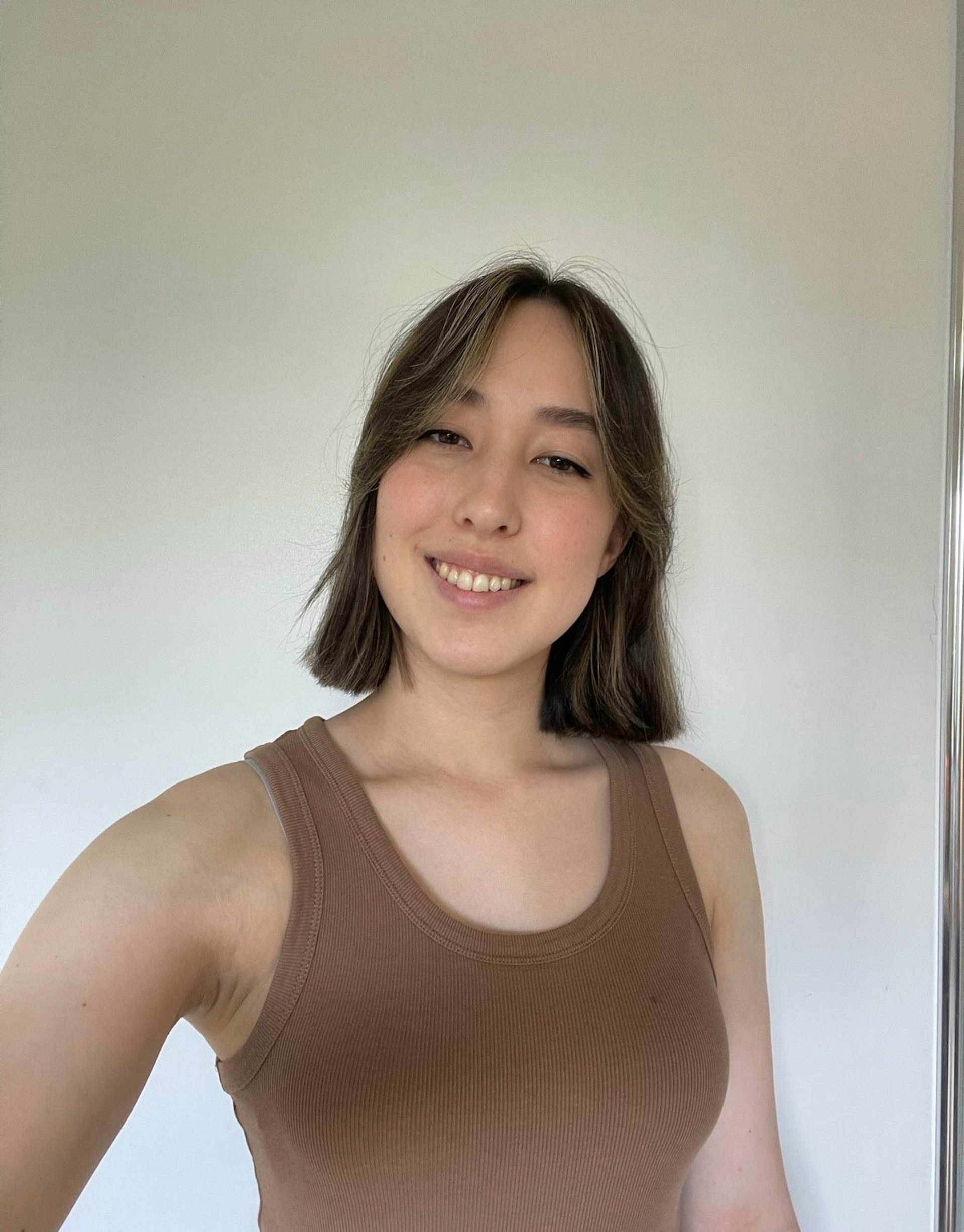 Lili stands taking a smiling selfie in front of white wall, wearing a brown singlet.