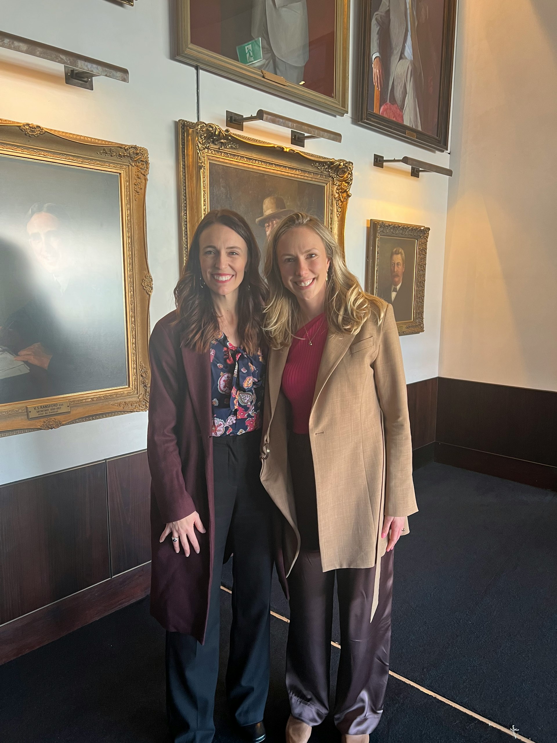 A photo of two women—Prime Minister Jacinda Ardern on the left, and Sharesies 3EO Brooke Roberts on the right. Both women are smiling into the camera, in front of a wall decorated with painted portraits. 