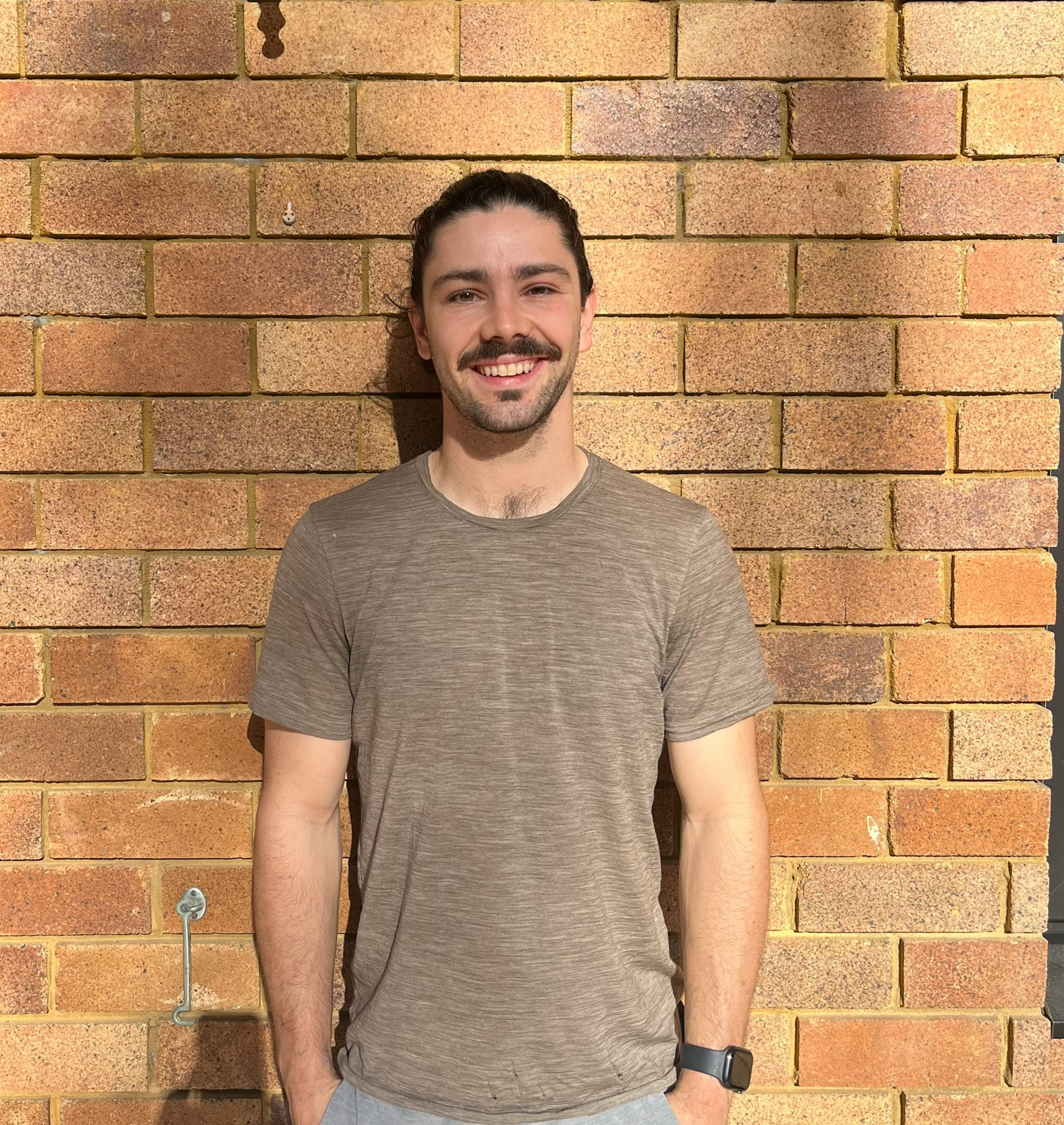 Tim stands in front of a brick wall, smiling at the camera with his hands in his pockets.