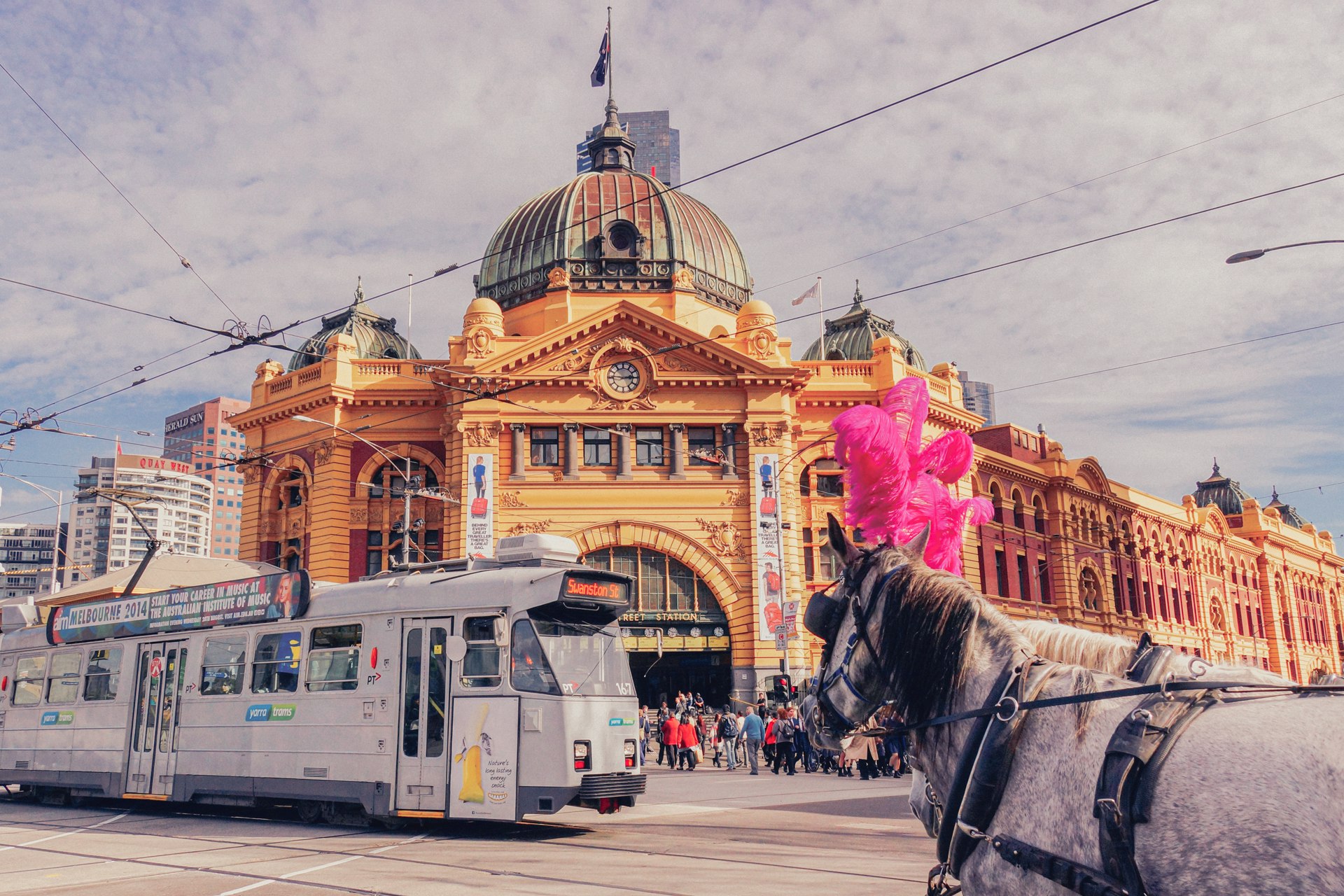 A busy day in Melbourne outside the iconic Flinders street station. A tram, as well as a horse and carriage, are getting people where they need to go.