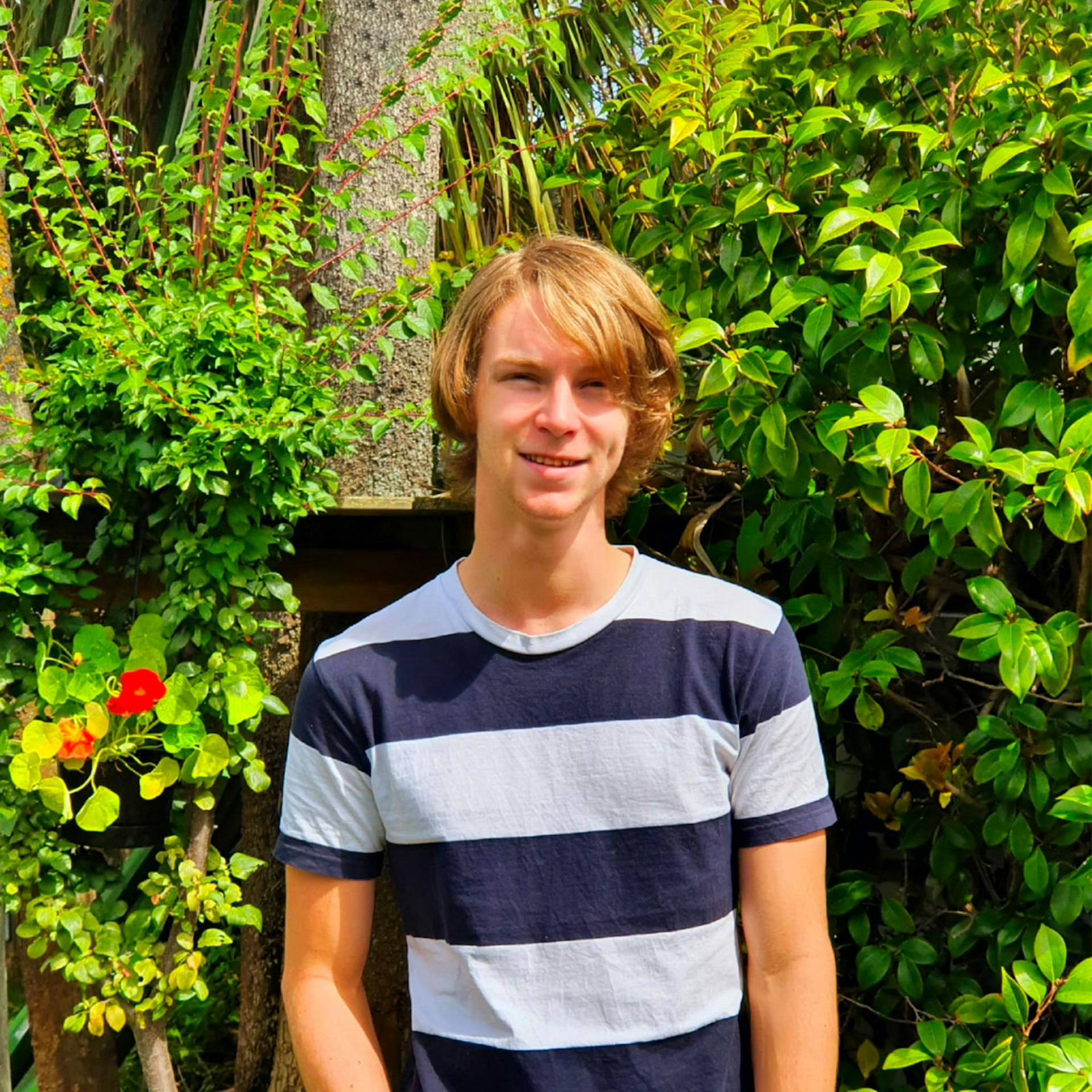 A young man wearing a striped t-shirt and jeans standing in front of some trees.