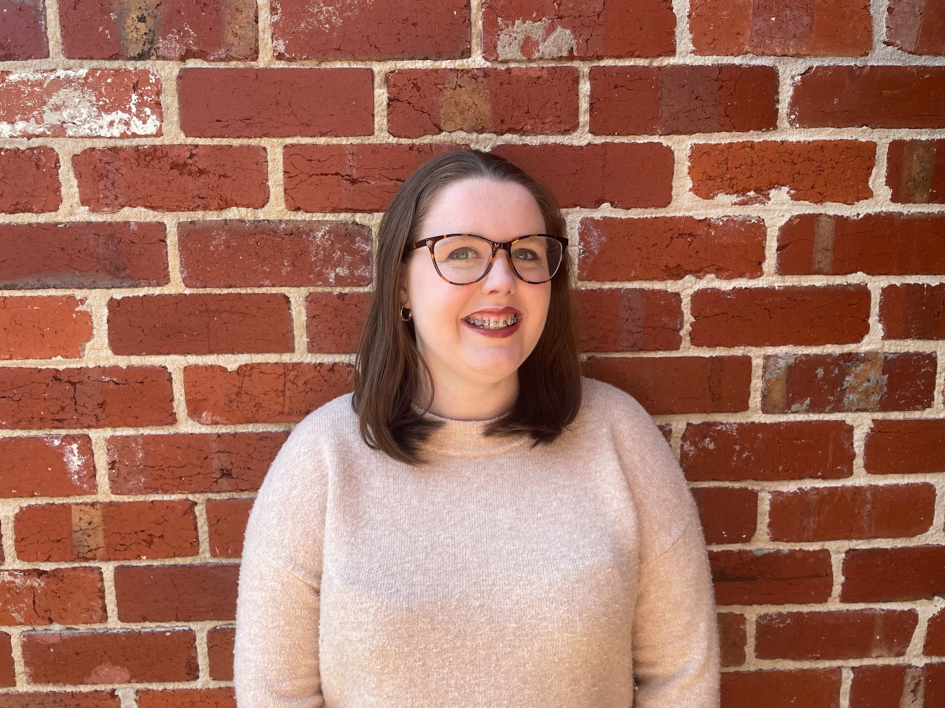 A woman wearing glasses smiling against a brick wall