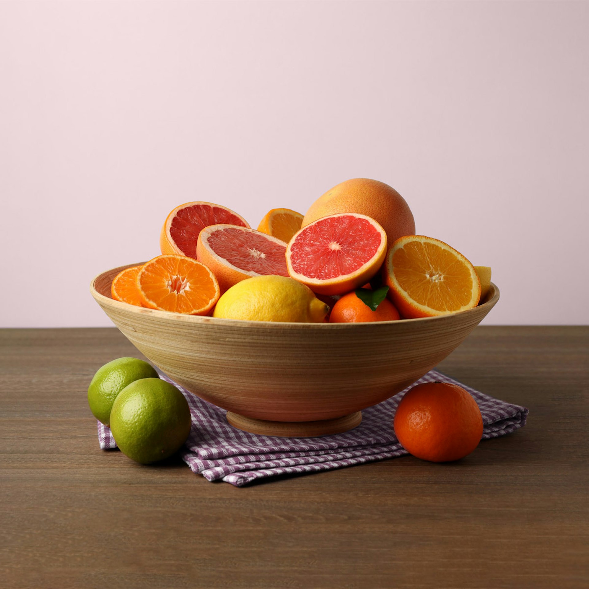 A wooden bowl of assorted citrus fruits is sitting on a tablecloth on top of a wooden table, in front of a light pink background.