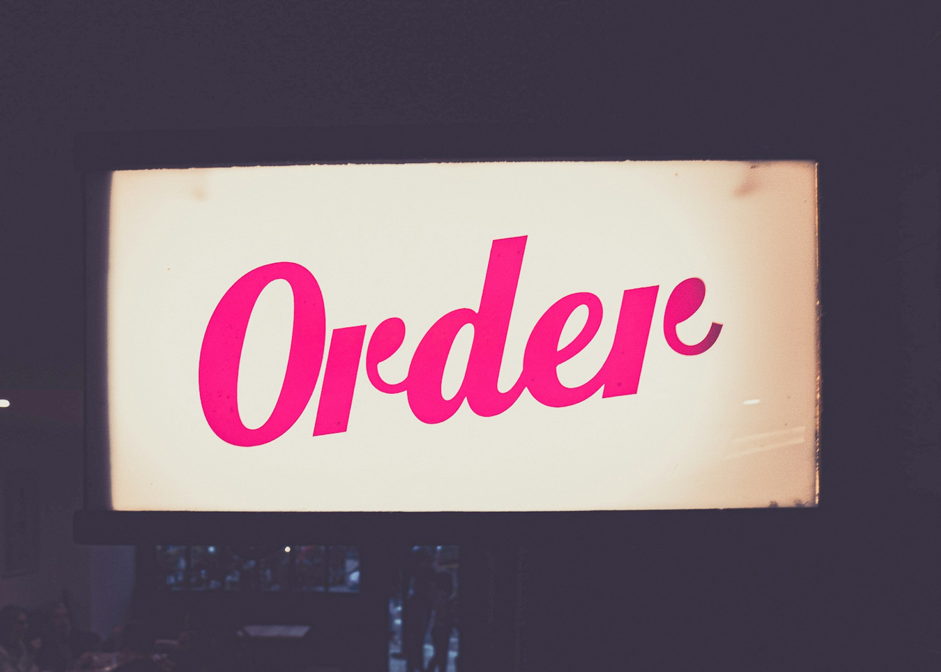 A white back-lit sign is sitting against a dark background, with the word 'Order' running across it diagonally (from bottom left to top right) in pink script. 