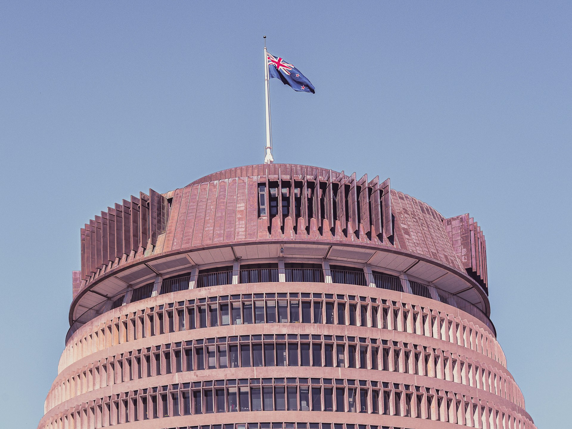 Top half of the Beehive building against a blue sky with the New Zealand flag flying.