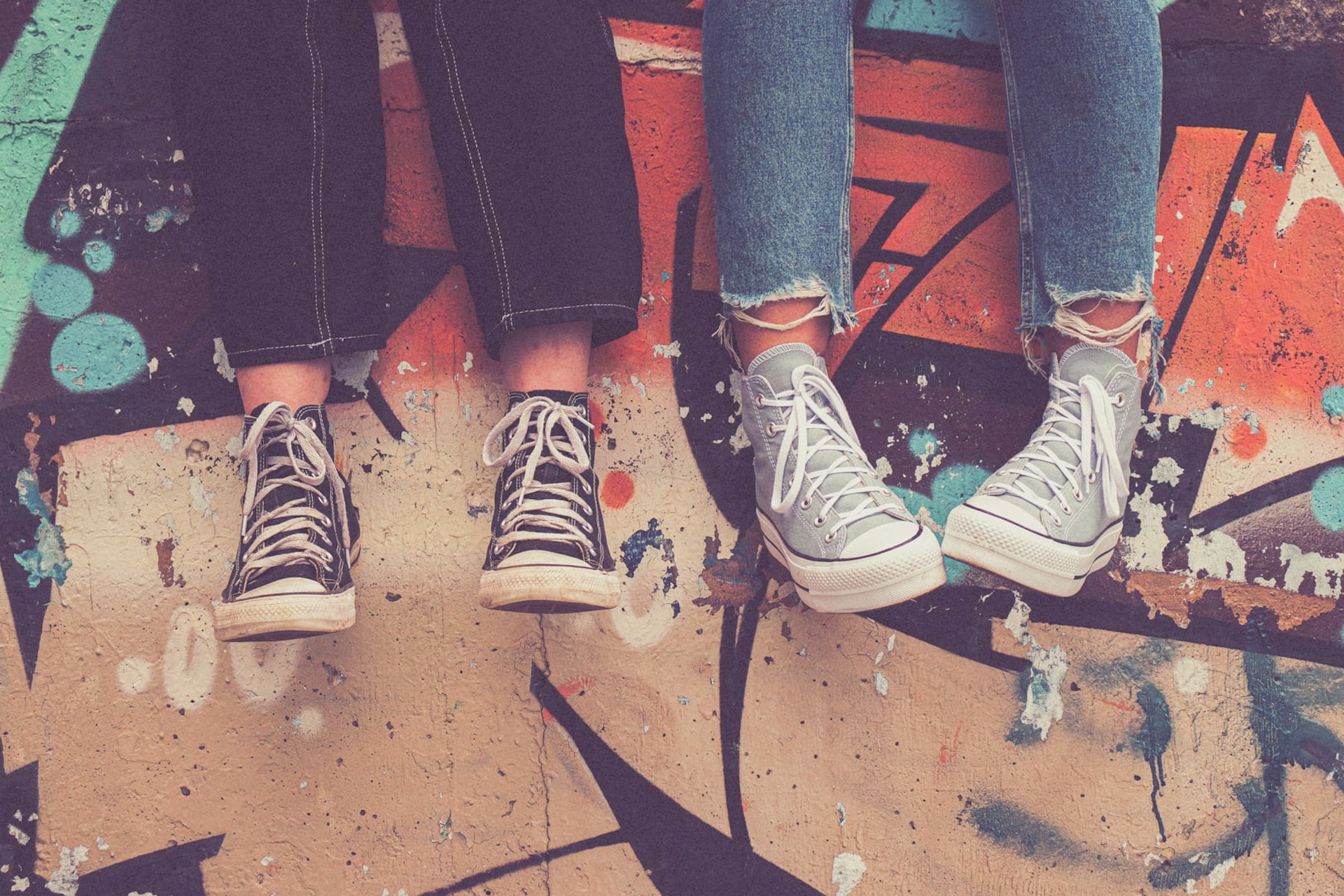Two pairs of feet wearing Converse sneakers are dangling against a graffiti-covered wall.