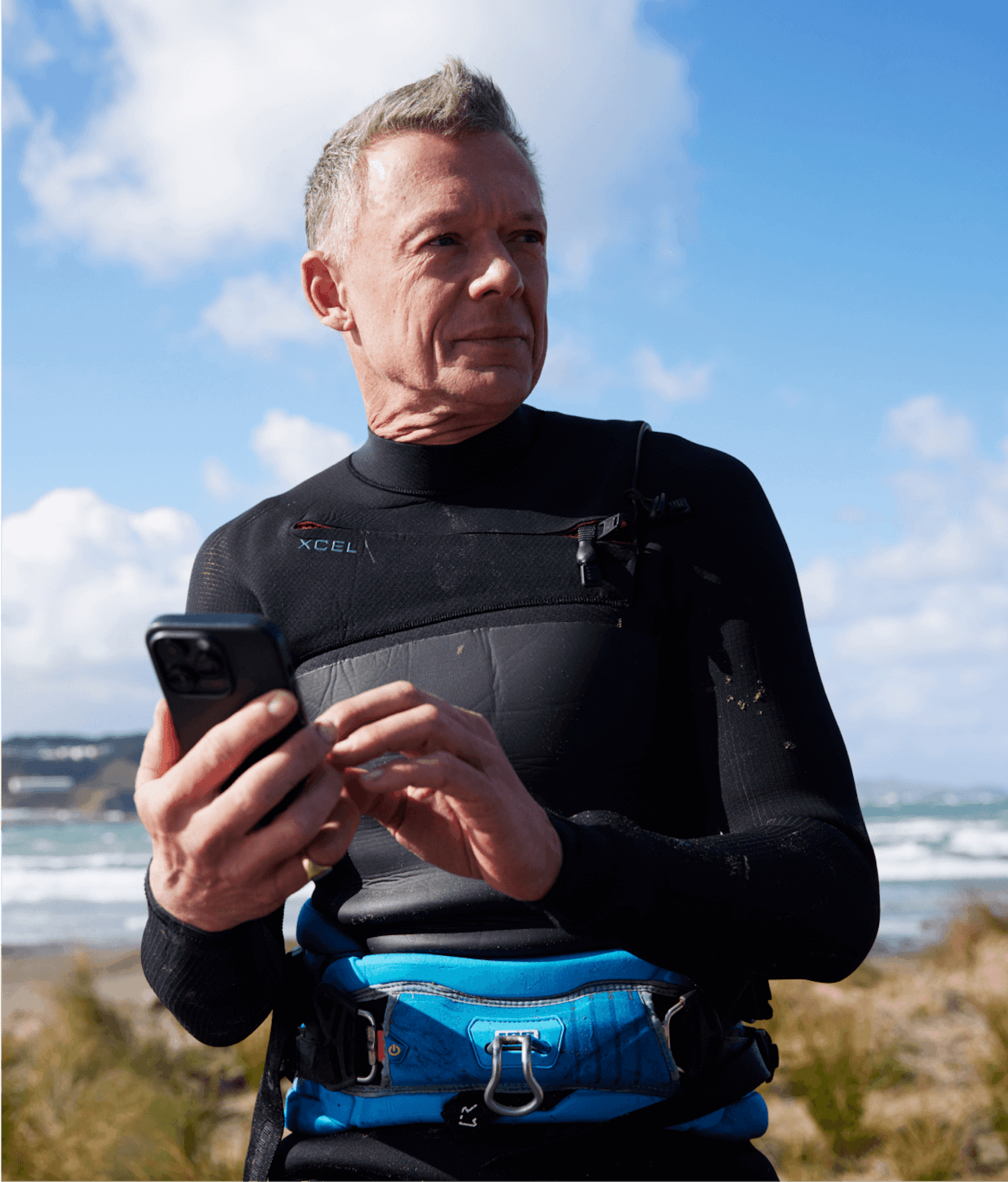 A middle-aged man in a wet suit holds a phone in his hand at the beach.