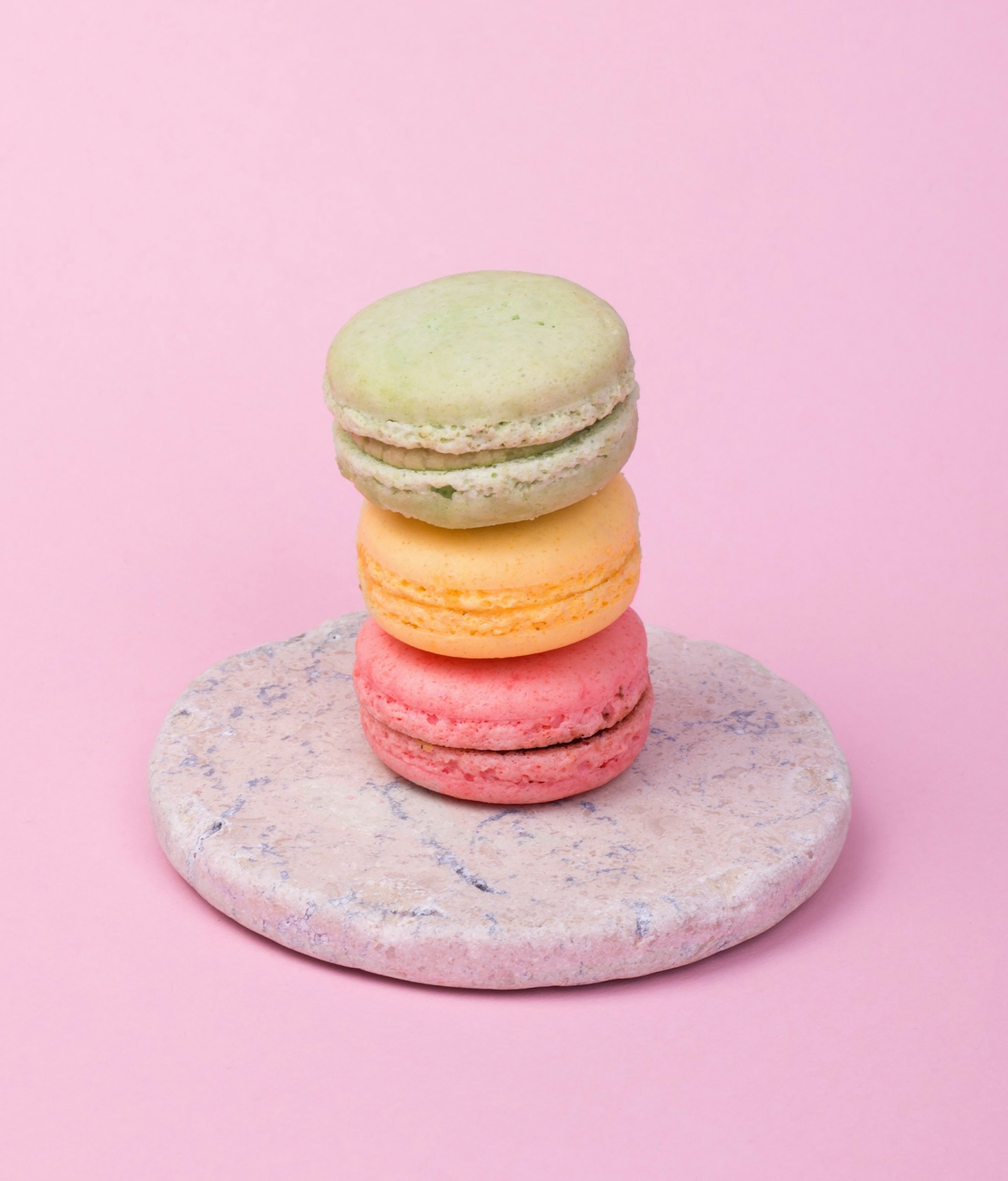 A green, orange, and pink macarons are stacked on top of each other, sitting on a plate in front of a pink background. 